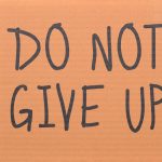 Cardboard sign reading 'Do Not Give Up' placed on green turf, symbolizing perseverance.
